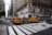 Yellow taxis are driving through a busy city intersection surrounded by tall buildings. A large department store is on the corner with a billboard ad above it. Pedestrians are crossing the street, and a traffic light is visible.