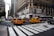 Yellow taxis are driving through a busy city intersection surrounded by tall buildings. A large department store is on the corner with a billboard ad above it. Pedestrians are crossing the street, and a traffic light is visible.