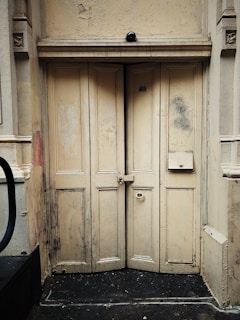 A timeworn, pale-colored wooden door with visible signs of aging and wear, such as stains, scuffs, and chipped paint. The door features multiple panels and a small, rectangular mailbox. An old surveillance camera is mounted above the door.