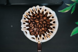 A top-down view of a coffee filter filled with dark roasted coffee beans. The filter is placed inside a dripper, surrounded by a few green leaves on a dark surface, creating a contrast between the coffee beans and the background, highlighting the texture of the beans and the filter.