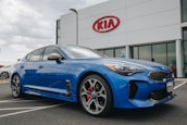 A modern blue Kia sedan is parked in front of a dealership with a large Kia sign on the building. The car is angled slightly to the side, showcasing its sleek design and shiny exterior. The setting appears to be a car lot with overcast weather.
