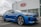 A modern blue Kia sedan is parked in front of a dealership with a large Kia sign on the building. The car is angled slightly to the side, showcasing its sleek design and shiny exterior. The setting appears to be a car lot with overcast weather.