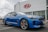 A modern blue Kia sedan is parked in front of a dealership with a large Kia sign on the building. The car is angled slightly to the side, showcasing its sleek design and shiny exterior. The setting appears to be a car lot with overcast weather.
