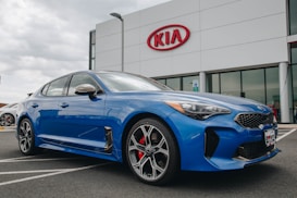 A modern blue Kia sedan is parked in front of a dealership with a large Kia sign on the building. The car is angled slightly to the side, showcasing its sleek design and shiny exterior. The setting appears to be a car lot with overcast weather.