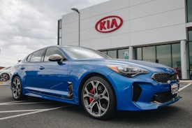 A modern blue Kia sedan is parked in front of a dealership with a large Kia sign on the building. The car is angled slightly to the side, showcasing its sleek design and shiny exterior. The setting appears to be a car lot with overcast weather.