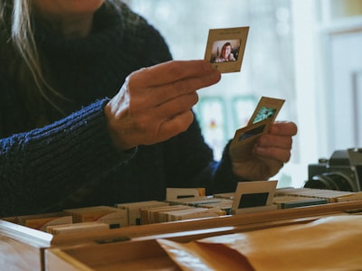 A person is holding photo slides close to a collection of similar slides in a wooden box. The scene is indoors and softly lit, suggesting an atmosphere of nostalgia. A camera is partially visible on the right side of the image.