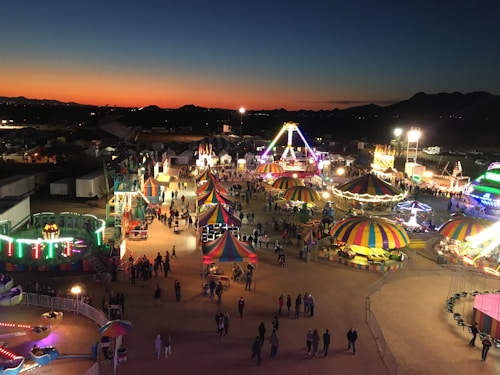 A lively fairground with various colorful tents and rides is bustling with people as the sun sets in the background. The sky displays a gradient of orange to deep blue, providing a picturesque backdrop. Amusement rides and booths are illuminated with vibrant lights, adding to the festive atmosphere.