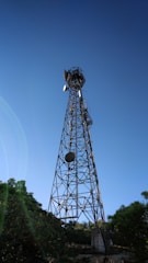 A tall metal communications tower stands against a clear blue sky, with a few satellite dishes attached to its structure. The tower is surrounded by lush green trees at its base, casting shadows on the ground.