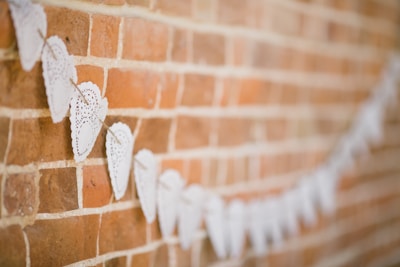 A string of white, heart-shaped paper doilies is hung along a rustic brick wall. The textured bricks provide a warm background that contrasts with the delicate, intricate lace design of the doilies.