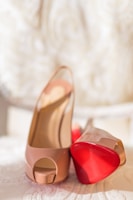 A pair of elegant high-heeled shoes with peep toes, featuring a glossy nude finish and distinctive red soles. The shoes are presented on a soft, textured surface with an intricate patterned background.