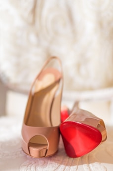 A pair of elegant high-heeled shoes with peep toes, featuring a glossy nude finish and distinctive red soles. The shoes are presented on a soft, textured surface with an intricate patterned background.
