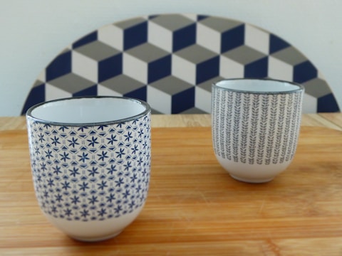 Two ceramic cups with intricate patterns are placed on a wooden surface. The cup in the foreground has a pattern of blue stars, while the cup in the background features a leafy design. A geometric pattern is visible in the backdrop.