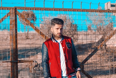 A young man is standing in front of a wire fence with barbed wire on top. He is wearing a red and black varsity jacket over a white shirt. The background shows a dry, possibly desert environment with trees and buildings in the distance under a bright blue sky.