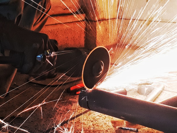 A person performing metal cutting with a circular saw, producing bright, intense sparks. The setting appears to be industrial, with concrete flooring and metal pipes.
