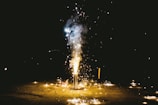 A firework is in full display at night, emanating bright sparks and smoke against a dark background. The ground is lit up with scattered sparkles, casting an ambient glow.