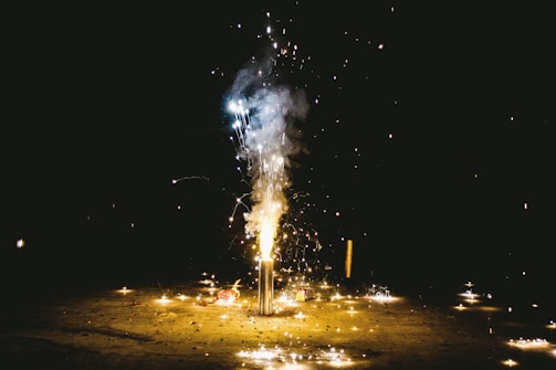 A firework is in full display at night, emanating bright sparks and smoke against a dark background. The ground is lit up with scattered sparkles, casting an ambient glow.
