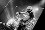 A musician on stage is energetically playing an electric guitar. Behind him, a drum set is visible amidst stage lights creating a lively atmosphere. The musician is wearing a distinctive hat with goggles, adding to his unique appearance. The scene is captured in black and white, emphasizing the dynamic movement and expressions.