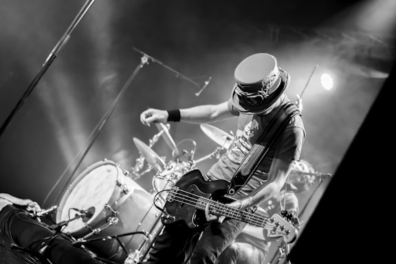 A musician on stage is energetically playing an electric guitar. Behind him, a drum set is visible amidst stage lights creating a lively atmosphere. The musician is wearing a distinctive hat with goggles, adding to his unique appearance. The scene is captured in black and white, emphasizing the dynamic movement and expressions.