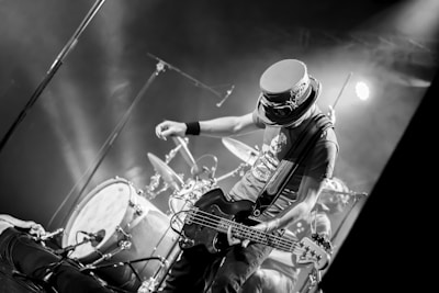 A musician on stage is energetically playing an electric guitar. Behind him, a drum set is visible amidst stage lights creating a lively atmosphere. The musician is wearing a distinctive hat with goggles, adding to his unique appearance. The scene is captured in black and white, emphasizing the dynamic movement and expressions.