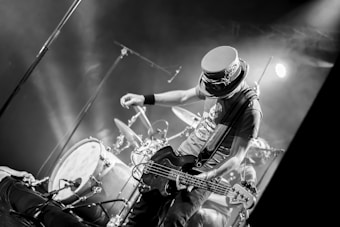 A musician on stage is energetically playing an electric guitar. Behind him, a drum set is visible amidst stage lights creating a lively atmosphere. The musician is wearing a distinctive hat with goggles, adding to his unique appearance. The scene is captured in black and white, emphasizing the dynamic movement and expressions.