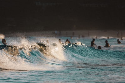 A group of people are swimming and surfing in the ocean, with waves cresting and sunlight reflecting off the water. The scene conveys a dynamic and lively atmosphere, as the movement of the waves is captured mid-action.