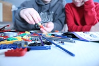 Two individuals are assembling various colorful plastic pieces and rods on a white surface, likely for a building or construction toy. One person is wearing a gray hoodie and is focusing on building, while the other, in a red shirt, observes closely. An instruction manual is open and visible on the table.