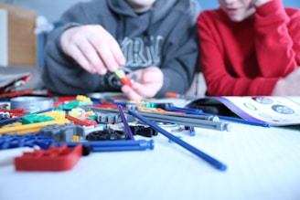 Two individuals are assembling various colorful plastic pieces and rods on a white surface, likely for a building or construction toy. One person is wearing a gray hoodie and is focusing on building, while the other, in a red shirt, observes closely. An instruction manual is open and visible on the table.
