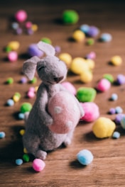 A soft toy bunny made of felt material is holding a pink polka-dotted egg. The bunny is placed on a wooden surface surrounded by colorful, fluffy pom-poms scattered around in various colors.