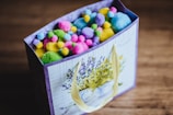 A decorated gift bag filled with colorful pom-poms in various pastel shades such as pink, yellow, blue, green, and purple. The bag has an elegant floral design featuring lavender and is adorned with cream handles.