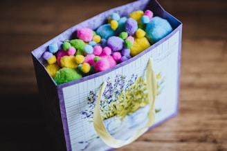 A decorated gift bag filled with colorful pom-poms in various pastel shades such as pink, yellow, blue, green, and purple. The bag has an elegant floral design featuring lavender and is adorned with cream handles.