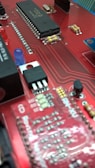 A close-up view of a red circuit board featuring various electronic components such as integrated circuits, resistors, capacitors, and a blue LED. The board has a network of intricate, printed connections and visible solder points.