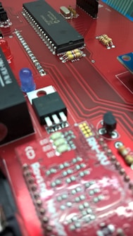 A close-up view of a red circuit board featuring various electronic components such as integrated circuits, resistors, capacitors, and a blue LED. The board has a network of intricate, printed connections and visible solder points.