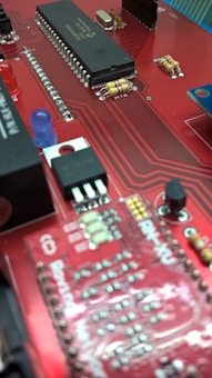 A close-up view of a red circuit board featuring various electronic components such as integrated circuits, resistors, capacitors, and a blue LED. The board has a network of intricate, printed connections and visible solder points.