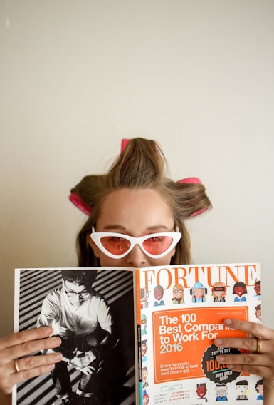 A person with hair rollers and wearing white cat-eye sunglasses is looking over a Fortune magazine. The magazine features a cover with an orange section titled 'The 100 Best Companies to Work For 2016'. The person holds the magazine while appearing to be in a relaxed setting.
