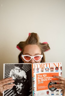 A person with hair rollers and wearing white cat-eye sunglasses is looking over a Fortune magazine. The magazine features a cover with an orange section titled 'The 100 Best Companies to Work For 2016'. The person holds the magazine while appearing to be in a relaxed setting.