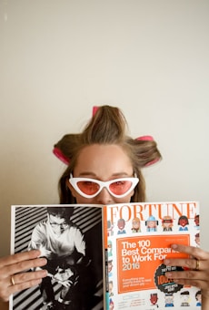 A person with hair rollers and wearing white cat-eye sunglasses is looking over a Fortune magazine. The magazine features a cover with an orange section titled 'The 100 Best Companies to Work For 2016'. The person holds the magazine while appearing to be in a relaxed setting.