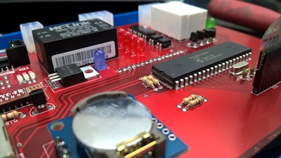 A close-up view of a red circuit board featuring various electronic components, including a microchip with multiple pins, capacitors, resistors, and an LED. The board also has a barcode sticker and a flat black component labeled 'HLK PXE04'. The intricate circuitry and small details are clearly visible.