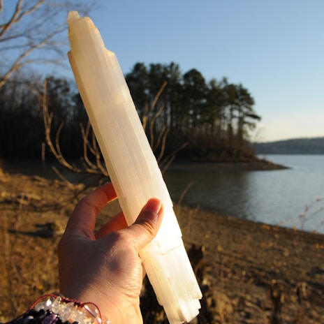 A hand holding a long, translucent crystal against the backdrop of a serene lake. The background shows a shoreline with sparse vegetation and a cluster of trees. The sunlight casts warm tones on the scene, highlighting the texture and clarity of the crystal.