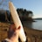 A hand holding a long, translucent crystal against the backdrop of a serene lake. The background shows a shoreline with sparse vegetation and a cluster of trees. The sunlight casts warm tones on the scene, highlighting the texture and clarity of the crystal.