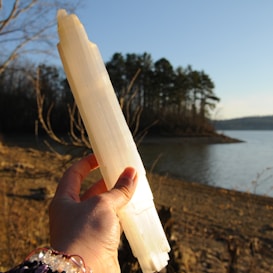 A hand holding a long, translucent crystal against the backdrop of a serene lake. The background shows a shoreline with sparse vegetation and a cluster of trees. The sunlight casts warm tones on the scene, highlighting the texture and clarity of the crystal.