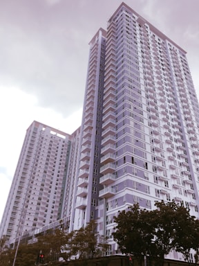 A pair of modern high-rise residential buildings with numerous balconies, large windows, and a sleek architectural design. The sky is overcast, and there are trees at the base of the buildings.