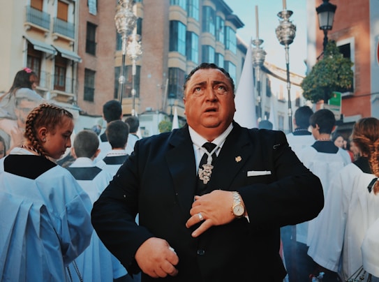 A man in a black suit, adorned with jewelry and a religious pendant, stands among a procession of people dressed in white liturgical robes. The background is an urban environment with buildings and street lamps, indicating a city street. People in the procession carry religious objects and focus appears to be on the solemn face of the man in the foreground.