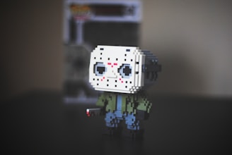 A pixelated figurine of a character wearing a white hockey mask with black eye holes, red markings, a green jacket, and blue pants. It stands on a dark surface with a blurry background featuring a boxed toy.