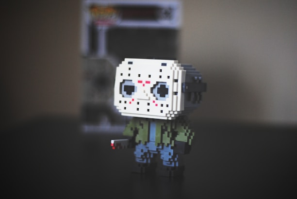 A pixelated figurine of a character wearing a white hockey mask with black eye holes, red markings, a green jacket, and blue pants. It stands on a dark surface with a blurry background featuring a boxed toy.