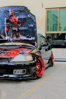 A customized black car with red rims and an open hood displaying a detailed graphic of a fictional character. The car appears to be parked in a tiled area next to a building with large windows.