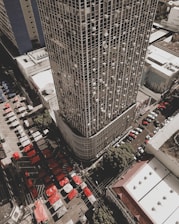 Aerial view of a densely populated urban area featuring a tall skyscraper with a grid pattern facade. Numerous air conditioning units are visible on the building's exterior. Below, a bustling market scene is unfolding with rows of colorful tents in red and white. Several streets are lined with parked cars, and the surrounding buildings have flat roofs and simplistic design.