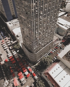 Aerial view of a densely populated urban area featuring a tall skyscraper with a grid pattern facade. Numerous air conditioning units are visible on the building's exterior. Below, a bustling market scene is unfolding with rows of colorful tents in red and white. Several streets are lined with parked cars, and the surrounding buildings have flat roofs and simplistic design.