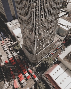 Aerial view of a densely populated urban area featuring a tall skyscraper with a grid pattern facade. Numerous air conditioning units are visible on the building's exterior. Below, a bustling market scene is unfolding with rows of colorful tents in red and white. Several streets are lined with parked cars, and the surrounding buildings have flat roofs and simplistic design.