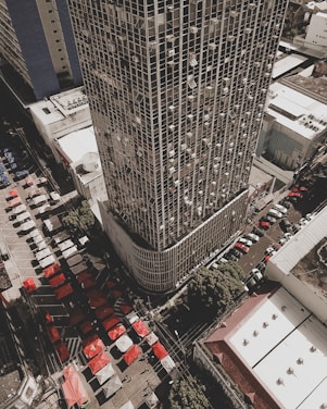 Aerial view of a densely populated urban area featuring a tall skyscraper with a grid pattern facade. Numerous air conditioning units are visible on the building's exterior. Below, a bustling market scene is unfolding with rows of colorful tents in red and white. Several streets are lined with parked cars, and the surrounding buildings have flat roofs and simplistic design.