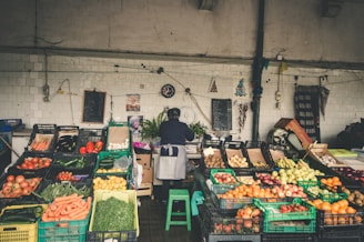 A market stall displaying a variety of fresh vegetables and fruits, including tomatoes, carrots, cucumbers, peppers, onions, and apples. The vendor, wearing an apron, stands behind the stall organizing the produce. The background consists of white tiled walls with hanging decorations, a black clock, and chalkboards.